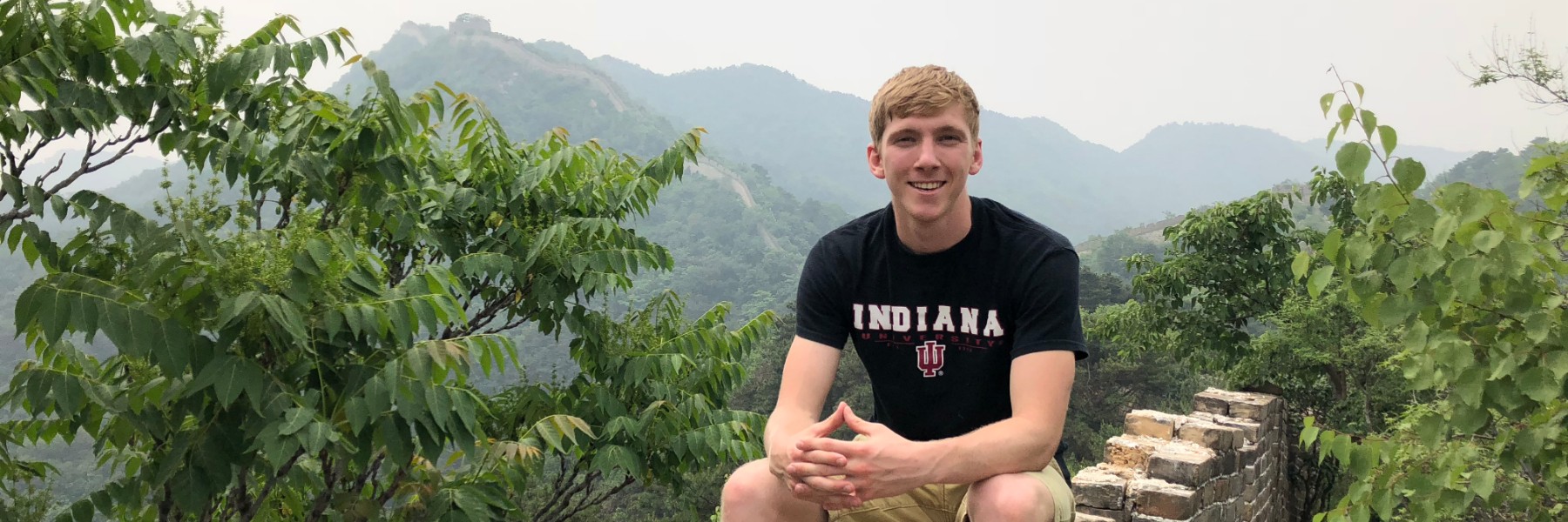 HIEP recipients in Asia and the Middle East - Calvin Isch poses for a photo while sitting on a section of the Great Wall of China with rolling hills and more wall behind him.