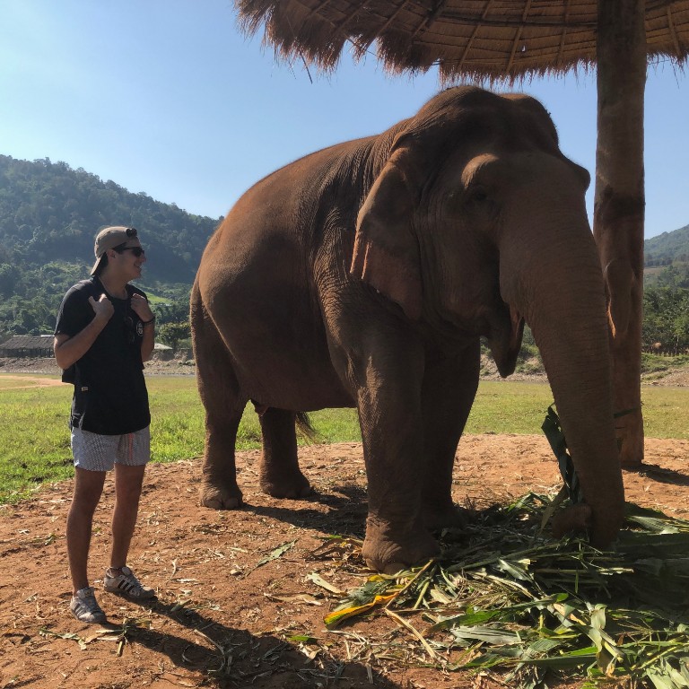 HIEP recipients in Asia and the Middle East - Christopher Kusovski stands in shorts next to an elephant in an open field with hills in the background. Photo taken near Chiang Mai in Thailand.