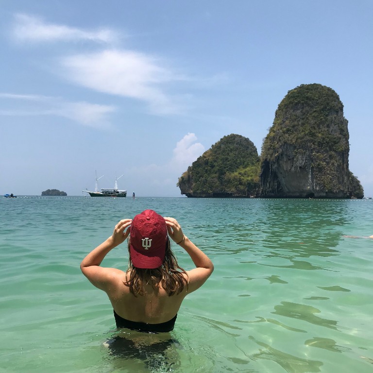 HIEP recipients in Asia and the Middle East - Riley Petty is swimming in the ocean near large rock outcrops. She faces away with an IU baseball cap turned toward the camera.