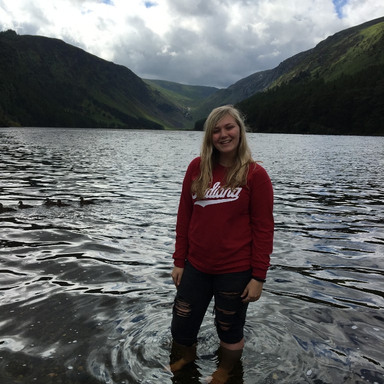HIEP students in the UK - Maya Painter poses in front of the Wicklow Mountains standing knee-deep in the upper lake of Glendalough, Ireland. Ducks swim by behind her.