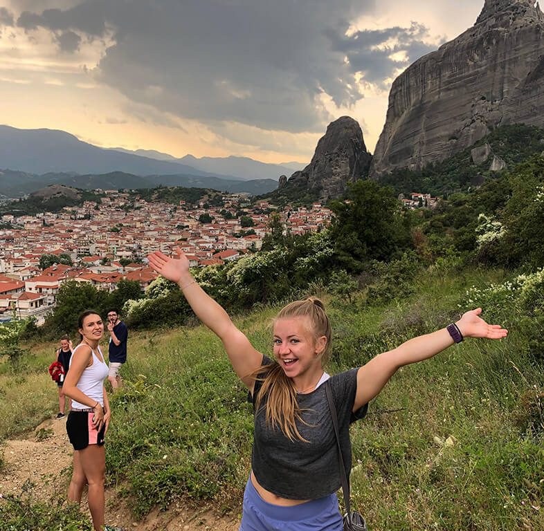 Wynne Milhouse poses with a smile and her arms in the air while on a hike outside Athens Greece in 2019